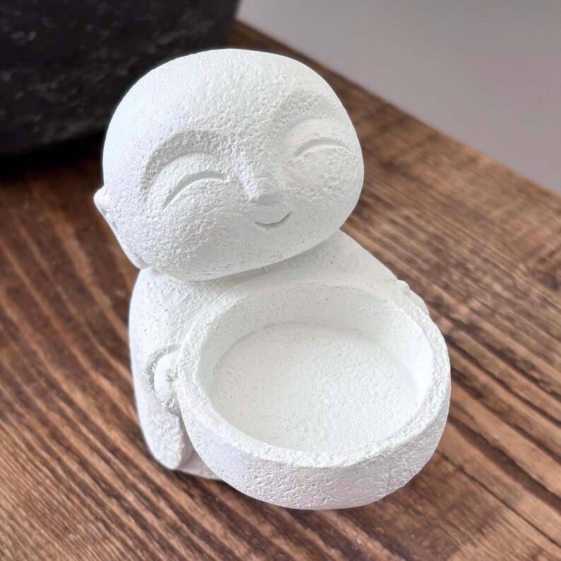 May include: A white, textured figurine of a smiling Buddha holding a small bowl. The figurine is made of a solid material and sits on a wooden surface. The Buddha's face has closed eyes and a gentle smile. The bowl is round and appears to be empty.
