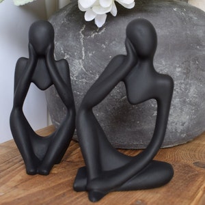 May include: Two black abstract ceramic sculptures of people sitting with their hands on their faces. The sculptures are on a wooden surface.