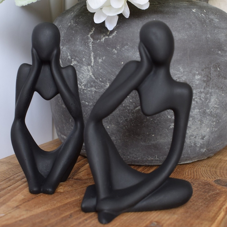 May include: Two black abstract ceramic sculptures of people sitting with their hands on their faces. The sculptures are on a wooden surface.