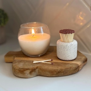 May include: A lit, white candle in a clear glass container sits on a wooden cutting board. A small, speckled, white container holds a bundle of matches with brown tips. Two matches are crossed on the board.
