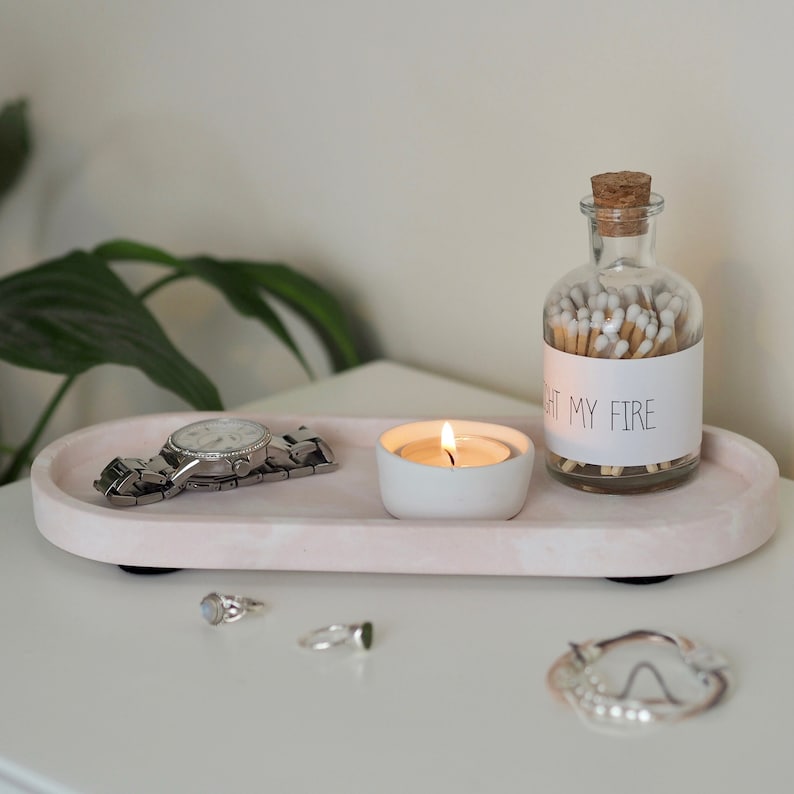 Puede incluir: Una bandeja ovalada rosa con un reloj, un peque&ntilde;o portavelas blanco con una vela encendida y un frasco de vidrio con cerillas que dice "Light My Fire".