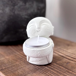 May include: A white, smiling Buddha figurine holding a lit tea light candle. The figurine has a textured surface and is set on a wooden surface. The candle is in a silver metal holder.