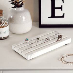 May include: White marble ring holder with five slots for rings. The holder has a white marble finish and is sitting on a white surface. There are five rings on the holder, each with a different gemstone.
