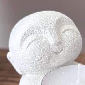 May include: A white, smiling Buddha statue with a textured surface. The statue is holding a white tea light candle. The Buddha's eyes are closed in a peaceful expression. The overall theme is one of serenity and meditation.