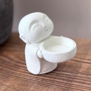 May include: A white, textured figurine of a smiling Buddha holding a small bowl. The figurine has a serene expression and is likely made of a plaster or ceramic material. The bowl is held out, suggesting it could be used as a small dish or candle holder.