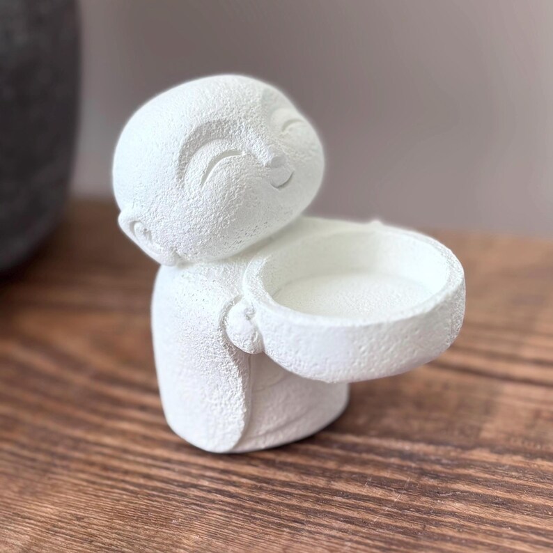 May include: A white, textured figurine of a smiling Buddha holding a small bowl. The figurine has a serene expression and is likely made of a plaster or ceramic material. The bowl is held out, suggesting it could be used as a small dish or candle holder.