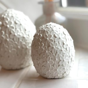 May include: Two white ceramic eggs with a floral design. The eggs are smooth and have a raised flower pattern.