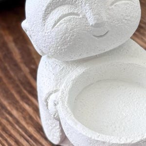 May include: A white, textured figurine of a smiling figure holding a small bowl. The figurine has a smooth, matte finish and is set against a blurred wooden background. The figure's face has a serene expression.
