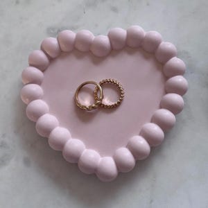 May include: A pink heart-shaped dish with a raised, beaded edge. Two gold rings are resting inside the dish.