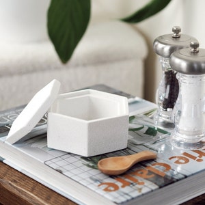 May include: A white hexagonal ceramic box with a lid, a wooden spoon, and two glass salt and pepper shakers with silver tops. The box and shakers are on a book with a grid pattern.