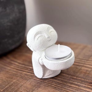 May include: A white, textured Buddha figurine holding a small, round candle holder with a lit white candle. The figurine has a smiling face and is made of a matte, possibly ceramic material. The background is blurred, with a wooden surface.