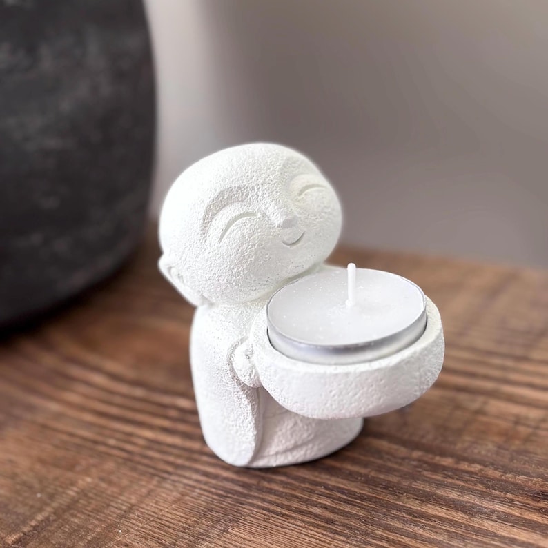May include: A white, textured Buddha figurine holding a small, round candle holder with a lit white candle. The figurine has a smiling face and is made of a matte, possibly ceramic material. The background is blurred, with a wooden surface.