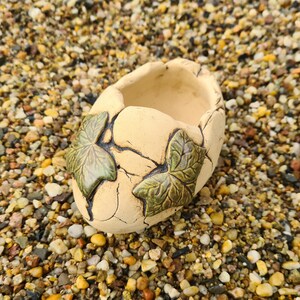 Könnte beinhalten: Ein kleiner, cremefarbener Keramiktopf mit einer rauen, strukturierten Oberfläche. Der Topf ist mit grünen Efeublättern verziert und hat eine abgerundete, leicht ovale Form.