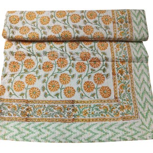 May include: A white cotton quilt with a floral pattern in shades of orange and green. The quilt has a border with a repeating pattern of flowers and leaves. The quilt is folded in half and the edges are visible.