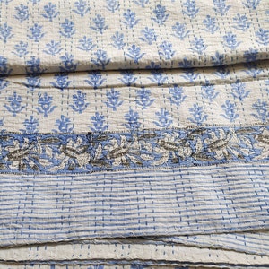May include: A white kantha quilt with a blue floral pattern and a blue and white striped border. The quilt is folded in thirds.