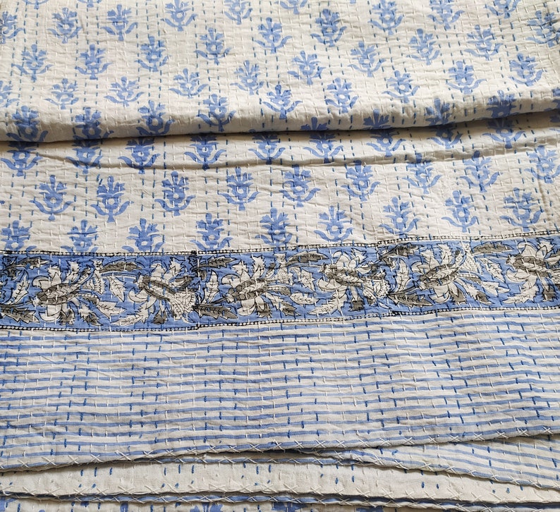 May include: A white kantha quilt with a blue floral pattern and a blue and white striped border. The quilt is folded in thirds.