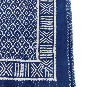 Op de afbeelding: Een close-up van een blauw en wit patroontextiel. Het ontwerp heeft een herhalend patroon van witte druppelvormen op een blauwe achtergrond. De rand heeft geometrische vormen en witte stiksels.