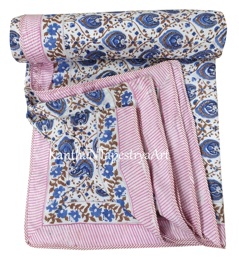 May include: A white cotton quilt with a blue and brown floral print. The quilt has a pink and white striped border with a brown stitched edge.