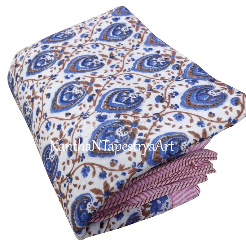 May include: A white cotton quilt with a blue and brown floral pattern. The quilt is folded in thirds and has a pink and white striped border.
