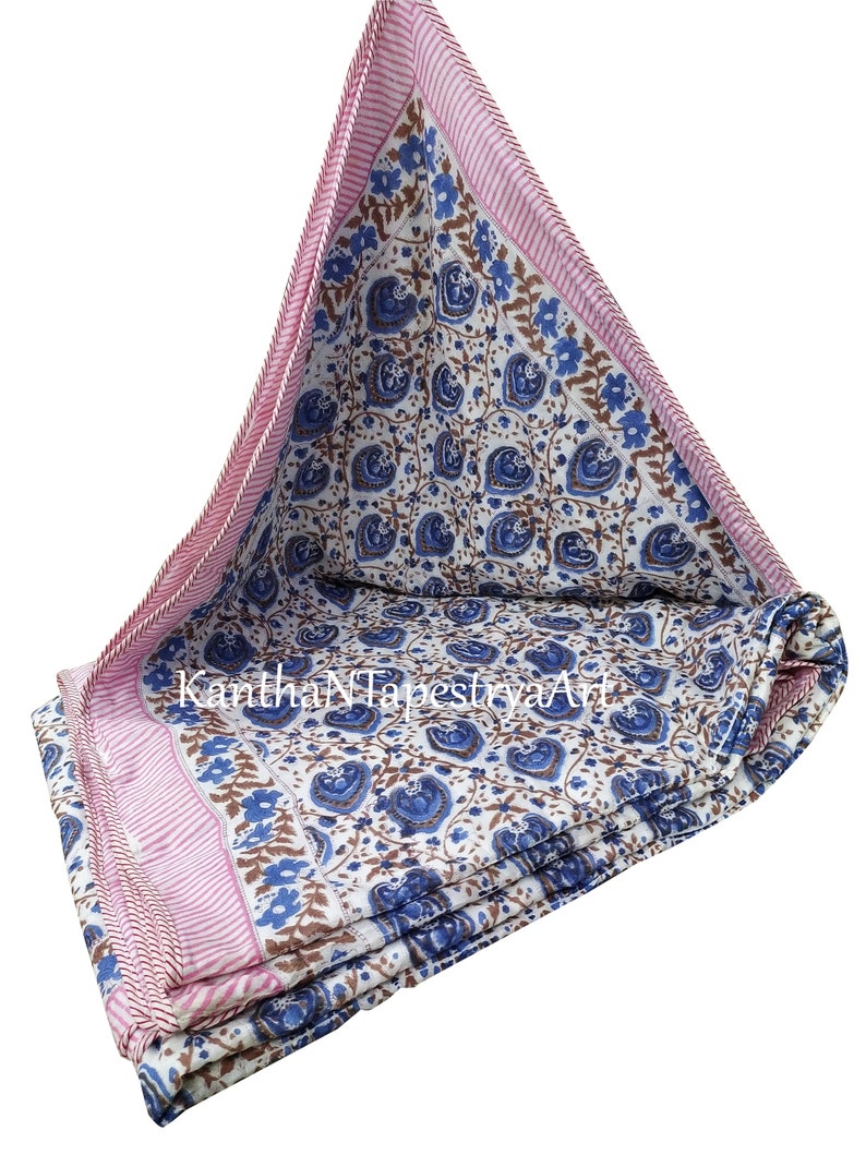 May include: A white cotton quilt with a blue and brown floral pattern. The quilt has a pink and white striped border. The quilt is folded in thirds and is lying on a white background.