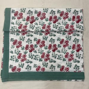 Puede incluir: Un tejido rectangular con fondo blanco y un patrón repetitivo de motivos florales circulares de color rojo oscuro y pequeñas ramas de hojas verdes. La tela está bordeada por una banda de color verde apagado sólido en los bordes izquierdo e inferior.
