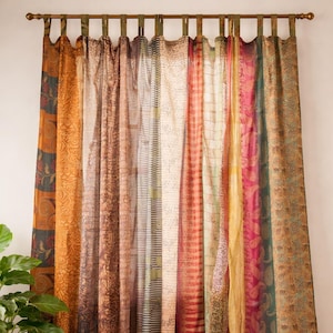 Peut inclure: Rideaux colorés et à motifs, suspendus à une tringle en bronze. Les rideaux présentent des tissus variés aux tons orange, marron, rose et vert. Les motifs incluent des motifs floraux, cachemire et rayés. Un pot de plante est visible à gauche.