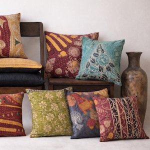 May include: An assortment of decorative throw pillows in various colors and patterns. Pillows feature floral, geometric, and patchwork designs in shades of red, green, blue, and gold. Some pillows are stacked on wooden chairs, while others are arranged on the floor.