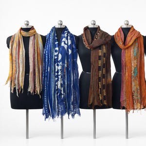 May include: Six long scarves displayed on black mannequins. The scarves feature varied patterns and colors including yellow, blue, brown, orange, teal, and pink. Each scarf has a lightweight, textured appearance with frayed edges and distinct prints.