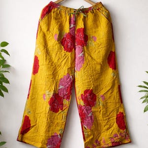 May include: Yellow wide-leg trousers with a red and pink floral pattern. The trousers are hanging on a wooden hanger against a white wall. The floral design is a repeating pattern of large roses.