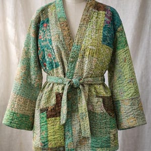 Może przedstawiać: Kurtka w stylu kimono z patchworku w odcieniach zieleni, turkusu i brązu. Kurtka ma dekolt w serek, długie rękawy i pas do wiązania. Tkanina wydaje się pikowana z widocznymi przeszyciami i wzorzystymi łatami.