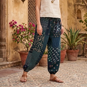 Può includere: Una canotta beige senza maniche abbinata a pantaloni patchwork color verde ottanio scuro con vari motivi floreali e geometrici, vita elasticizzata con coulisse e caviglie arricciate, indossati con sandali marroni e una collana di perline.