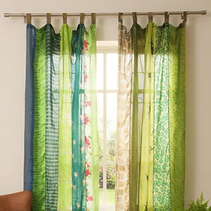 May include: A pair of vibrant, patchwork curtains with vertical panels in green, blue, and brown. The panels display floral and geometric patterns. The curtains are hung in front of a window, with a brown leather armchair and a small wooden table with a plant in the foreground.