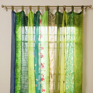 May include: A set of colorful, patchwork curtains in shades of green, blue, and beige. The curtains are made of various fabrics with floral, striped, and patterned designs. The curtains hang from a metal rod, and the sunlight filters through the fabric.