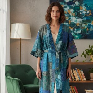 May include: A patchwork robe in shades of blue, teal, and grey, with a matching belt. The robe has a V-neck and short sleeves. The garment is worn over a cream-coloured dress. The background includes a green chair, a lamp, and a painting.