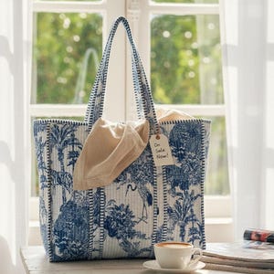 May include: A blue and white tote bag with a toile pattern featuring trees, birds, and tigers. The bag has a matching handle and a beige pouch. A tag reads "On Sale Now!" A cup of coffee sits on a table.