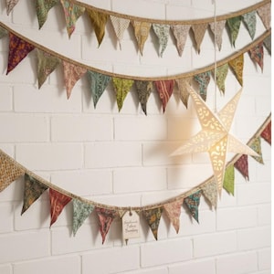 Puede incluir: Tres hileras de banderines de tela colorida cuelgan contra una pared de ladrillos blancos, con una linterna de papel en forma de estrella. Los banderines presentan banderas triangulares en varios patrones y colores. Una pila de tela está sobre una mesa de madera.