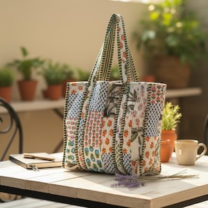 以下が含まれることがあります： 青、オレンジ、白の色合いの、花柄と幾何学模様のパッチワークトートバッグ。バッグには緑のストライプのハンドルとトリムが付いています。バッグは、ノート、ペン、カップが置かれた木製のテーブルの上にあります。