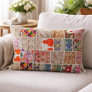 Puede incluir: Cojín rectangular de patchwork con una variedad de cuadrados de tela coloridos. Los cuadrados presentan patrones florales, paisley y geométricos en tonos naranja, rosa, verde y azul. El cojín está sobre un sofá blanco.