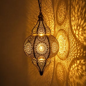 Può includere: Una lampada a sospensione con corpo metallico a forma di cipolla, caratterizzata da intricati motivi geometrici tagliati al laser. La lampada emette una calda luce dorata, proiettando ombre circolari dettagliate sulla parete adiacente.