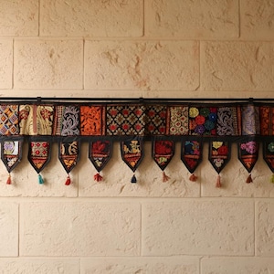 Op de afbeelding: Een decoratieve wandhanger met een zwarte stang en een reeks kleurrijke, stoffen panelen met patronen. Elk paneel heeft een uniek ontwerp in tinten rood, oranje en blauw. De onderkant heeft decoratieve kwastjes.