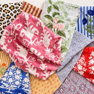 May include: A selection of folded fabric pieces featuring diverse patterns. Designs include leopard print, floral, geometric, and botanical motifs in shades of pink, blue, red, yellow, and white. The fabrics appear to be cotton or linen.