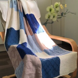 Può includere: Una coperta patchwork lavorata a maglia, drappeggiata su una sedia di legno. Presenta quadrati strutturati nei toni del blu, crema e beige. Un elemento d'arredo per la casa.