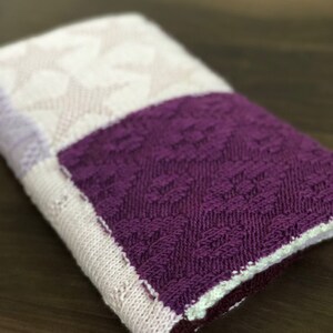 Può includere: Coperta di maglia piegata con un quadrato viola strutturato, una sezione bianca e lilla chiaro a fantasia e un bordo bianco. La coperta è realizzata in filato morbido ed è piegata con cura.
