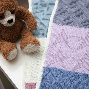 Può includere: Una coperta per neonati lavorata a maglia multicolore con un orsacchiotto. La coperta presenta sezioni in azzurro, lavanda e bianco, con motivi strutturati. L'orsacchiotto è marrone con muso e zampe bianche.