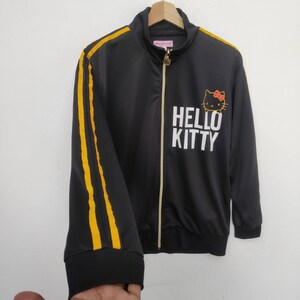 May include: Black zippered jacket with yellow stripes down the sleeves. The jacket has a Hello Kitty graphic with the text "Hello Kitty" in white.