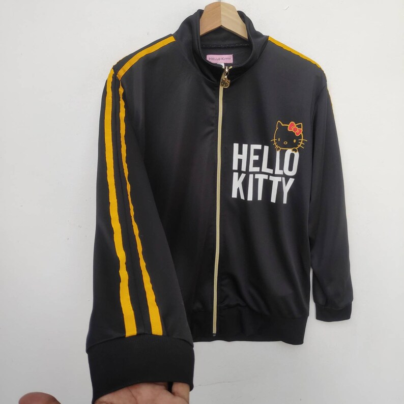 May include: Black zippered jacket with yellow stripes down the sleeves. The jacket has a Hello Kitty graphic with the text "Hello Kitty" in white.