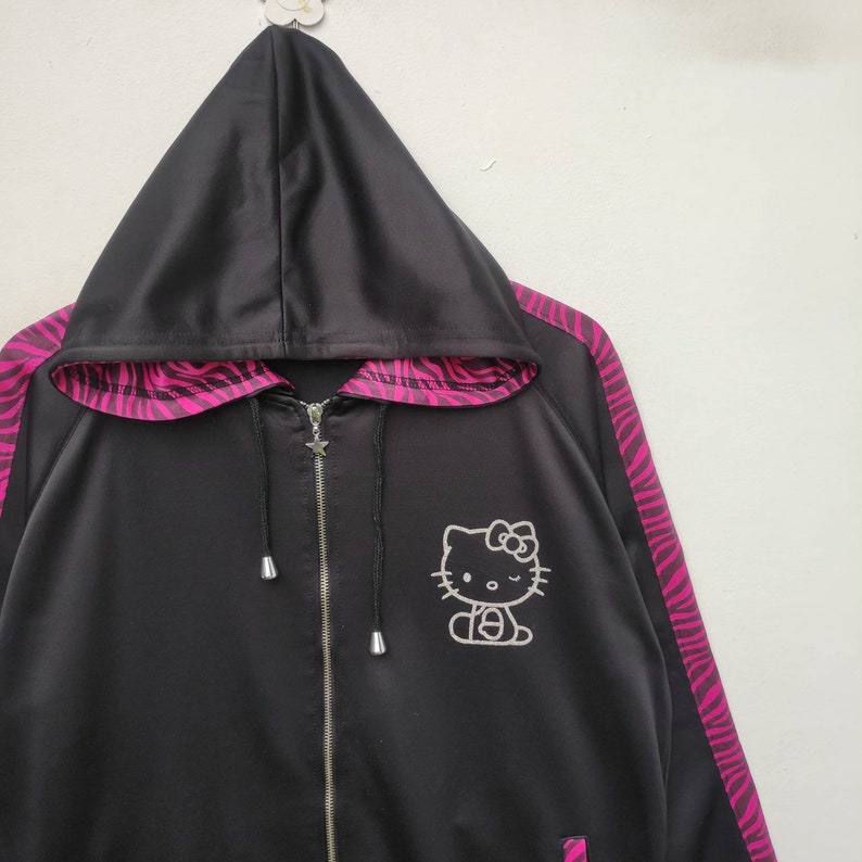 May include: Black satin hooded jacket with pink and black zebra stripes on the sleeves. The jacket has a zipper closure and a Hello Kitty graphic on the front.