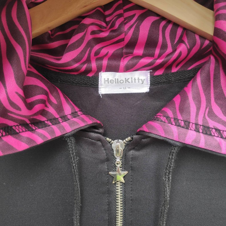 May include: Black zippered hoodie with a pink and black zebra print lining on the hood. The hoodie has a silver star zipper pull and a Hello Kitty label.