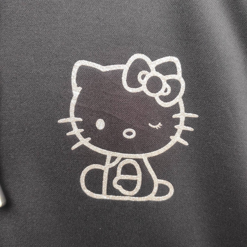 May include: A silver glitter Hello Kitty design on a black background. The design features Hello Kitty with a bow and winking.