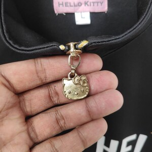 May include: Black zipper pull with a gold Hello Kitty charm. The charm features Hello Kitty's face with a red bow.